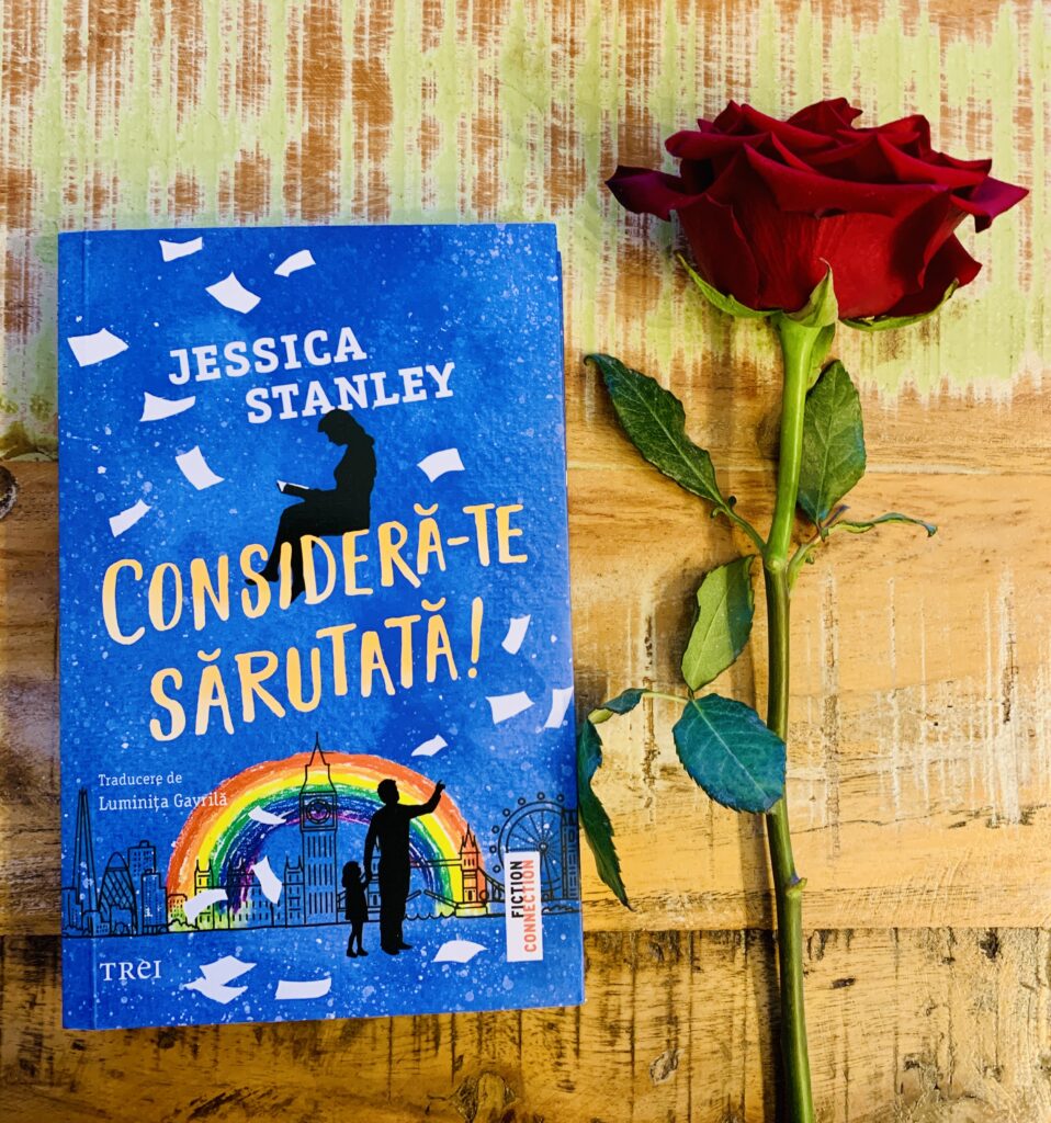 Romanul Consideră-te sărutată de Jessica Stanley așezat pe o masă de lemn rustică, lângă un trandafir roșu proaspăt, sugestie de mărțișor literar.