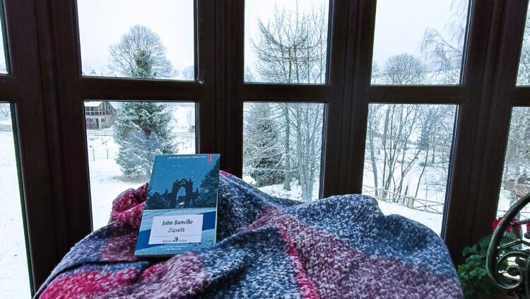 Coperta cărții „Zăpada” de John Banville, pe o pătură tricotată lângă o fereastră cu vedere la un peisaj înzăpezit.