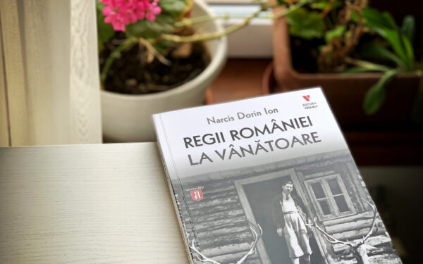 Coperta cărții Regii României la vânătoare așezată pe o masă, lângă o fereastră cu flori.