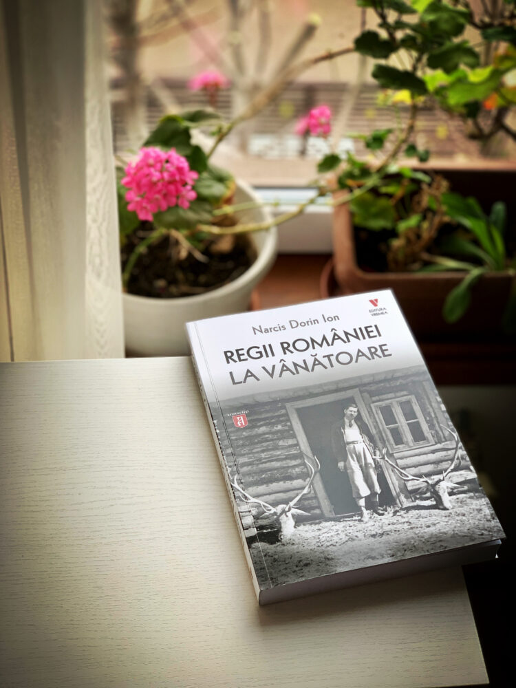 Coperta cărții Regii României la vânătoare așezată pe o masă, lângă o fereastră cu flori.