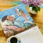 Cadru de relaxare cu o ceașcă de cafea, liliac mov și calendarul cu pictura Femeia care citește de Lovis Corinth