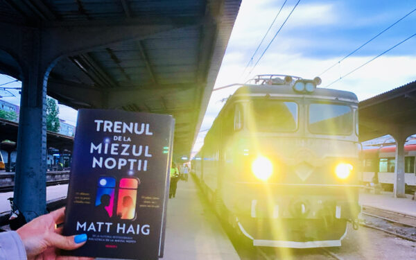 Noul roman Trenul de la miezul noptii de Matt Haig tinut in mana pe peronul garii langa o locomotiva de tren