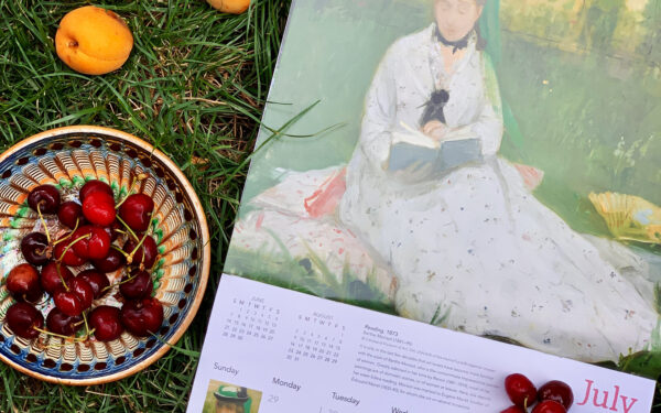 Calendar de perete cu tabloul Lectura de Berthe Morisot așezat pe iarbă verde lângă un bol cu cireșe și caise proaspete de vară.