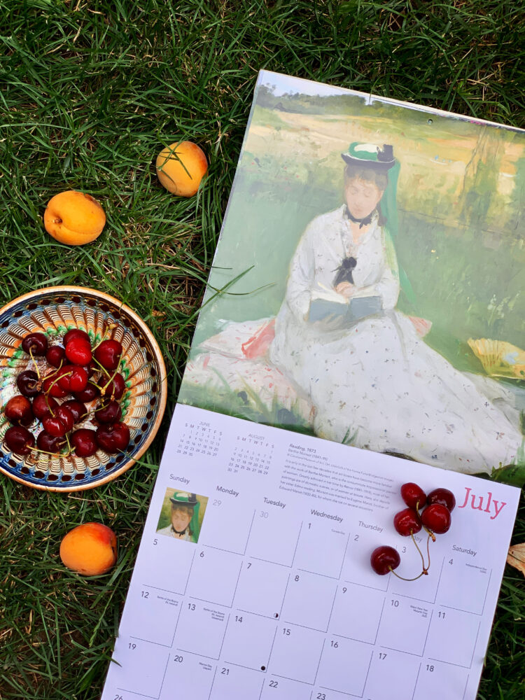 Calendar de perete cu tabloul Lectura de Berthe Morisot așezat pe iarbă verde lângă un bol cu cireșe și caise proaspete de vară.