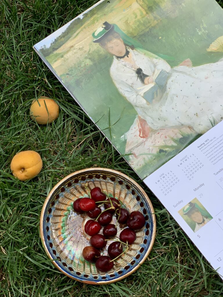 Detaliu de aproape cu pictura „Lectura” de Berthe Morisot într-un calendar de perete, așezat pe iarbă alături de o farfurie din ceramică plină cu cireșe și caise