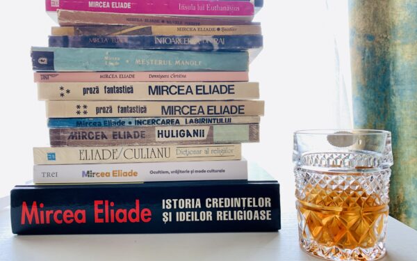 O stivă de cărți scrise de Mircea Eliade, printre care „Istoria credințelor și ideilor religioase” și „Huliganii”, așezată pe un birou lângă un pahar de whisky din cristal cu gheață.