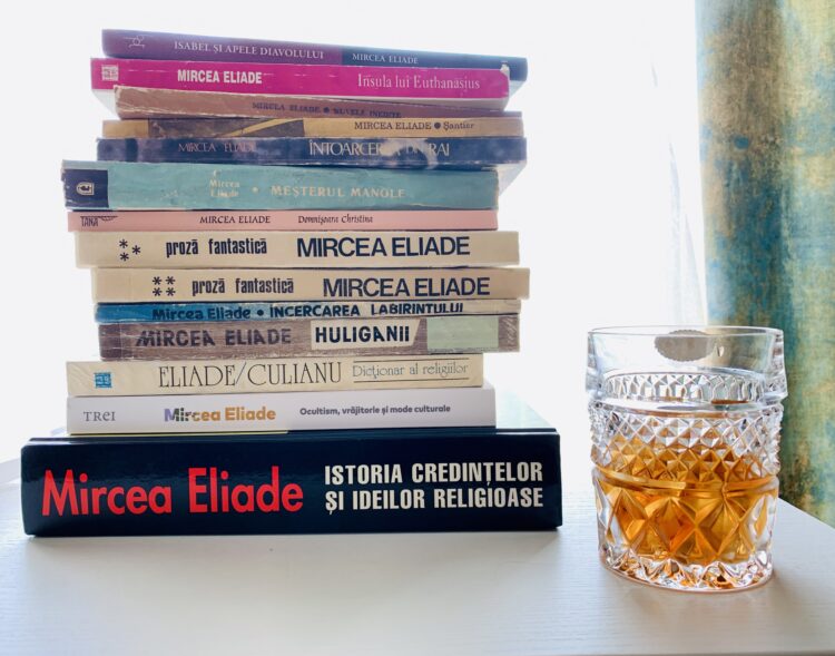 O stivă de cărți scrise de Mircea Eliade, printre care „Istoria credințelor și ideilor religioase” și „Huliganii”, așezată pe un birou lângă un pahar de whisky din cristal cu gheață.