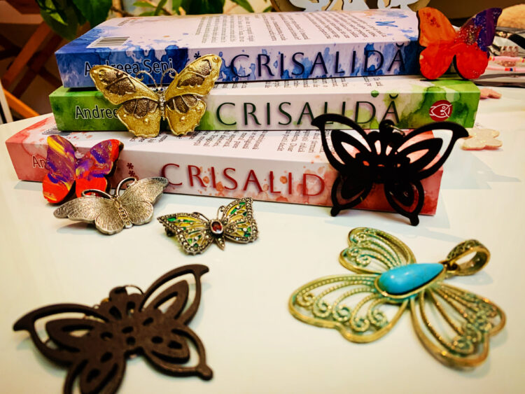 Cele trei volume ale Trilogiei Crisalida de Andreea Sepi, așezate unele peste altele și înconjurate de diverse decorațiuni, broșe și pandantive în formă de fluturi colorați.