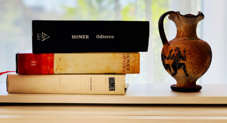Trei ediții diferite ale cărții Odiseea de Homer, stivuite orizontal pe o suprafață albă, alături de o vază ceramică în stil grecesc antic, pictată cu figuri negre pe fundal maro. Volumul de sus are o copertă neagră modernă de la Editura Art, cel din mijloc are o copertă portocalie, iar cel de jos este o ediție vintage de la Editura Univers.